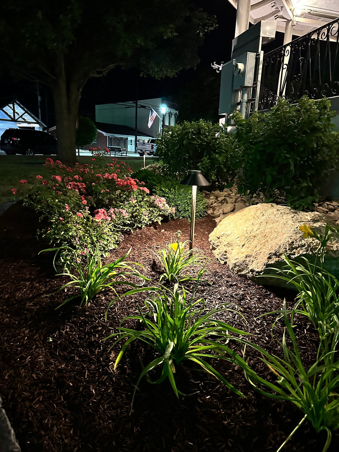 Nighttime view of a landscaped garden illuminated by a stylish pathway light, surrounded by vibrant flowers, greenery, boulders and mulch.