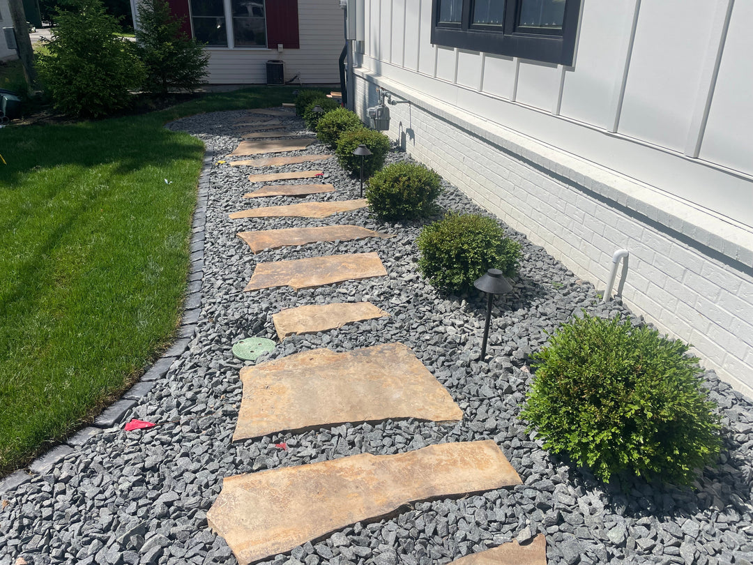 Stone pathway with natural flagstone steps surrounded by gray gravel and neatly trimmed shrubs, leading along the side of a house with modern landscaping.