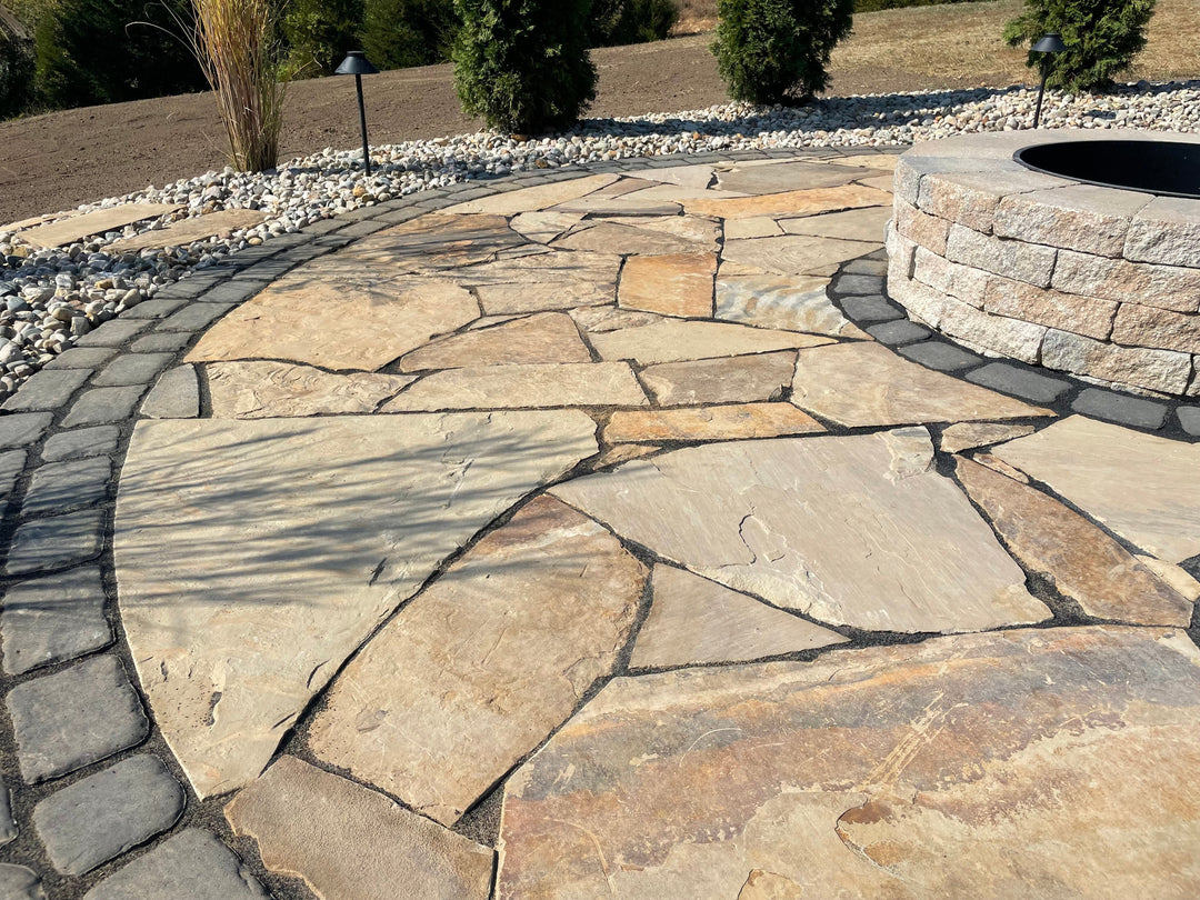 Outdoor patio featuring a natural flagstone surface with a fire pit surrounded by decorative stone edging, gravel accents, and lush landscaping in the background.