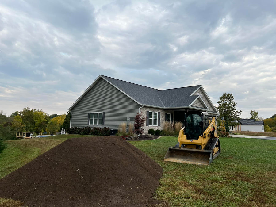 Large pile of screened topsoil on a green lawn beside a modern house, with a skid steer loader positioned nearby for landscaping work.