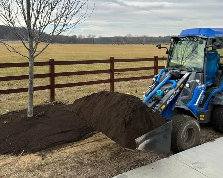 Blue loader transporting screened topsoil to a tree planting area next to a wooden fence, with an open field and cloudy sky in the background.